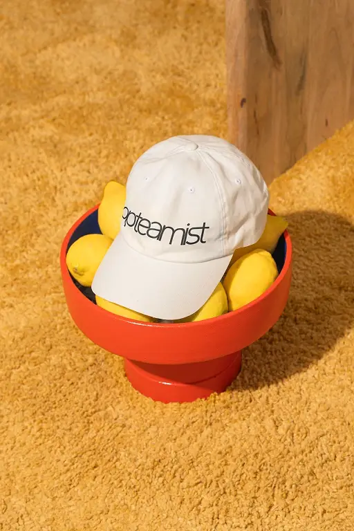 Gorra blanca con el wordmark de Opteamist bordado, dentro de un tazón rojo junto a limones