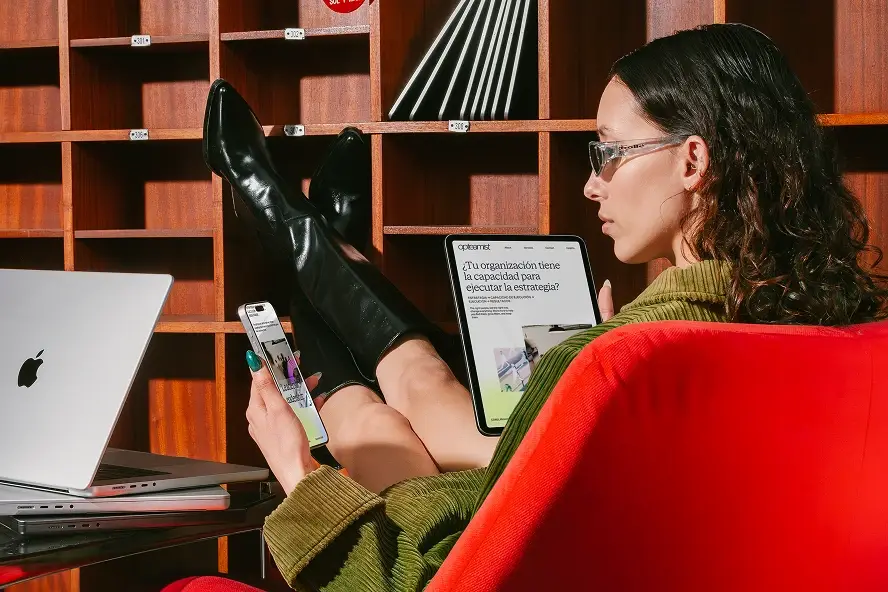 Una persona recostada en un sillón rojo revisa el sitio web de Opteamist en una tablet, con una laptop y un teléfono también mostrando el sitio